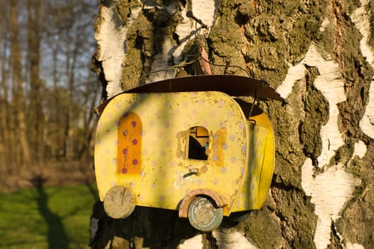 A yellow birdhouse in the shape of a caravan, attached to a tree trunk with white bark, in the sunlight. Ideal for nature and garden lovers.