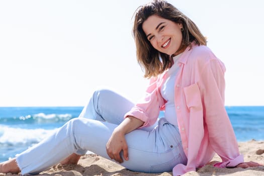 Young caucasian woman on vacation relaxing by the sea, sitting on the beach with a smile. Dressed in casual clothes, enjoying the sunny weather, ocean breeze, and peaceful summer atmosphere.
