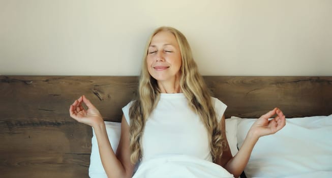 Calm relaxed healthy young woman meditates, girl practicing doing yoga exercises sitting in lotus pose on white bed at home in the morning, stress relief