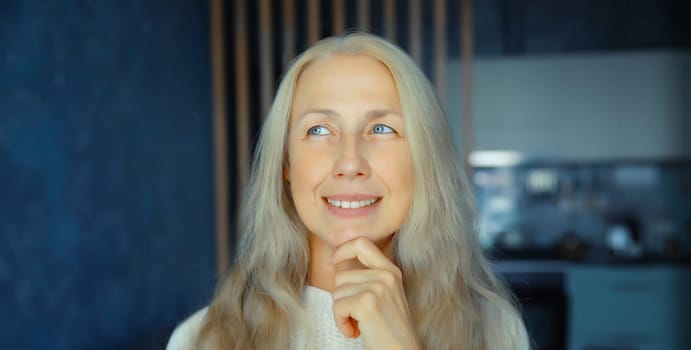 Pensive mature woman thinks looking out the window in early morning in living room with kitchen at home, copy space background