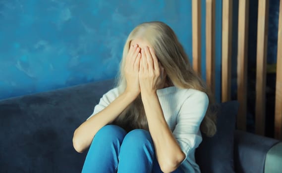 Upset sad crying woman covers her eyes and face with her hands while experiencing depression, stress, fatigue, mental suffering sitting on the couch at home