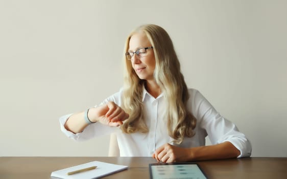 Confident mature woman professional working on digital tablet computer with paper documents looks at watch, checks the time, sitting at desk in office or at home