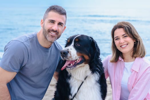 Portrait of a smiling caucasian couple with their large dog on the beach during vacation. Relaxed and joyful moment by the sea, with all three looking at the camera on a bright day.