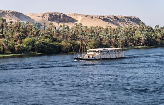 Dahabiya Nile cruise ship sailing alongside a sandy desert with palm trees on the River Nile near Aswan