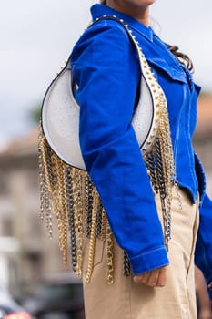Milan, Italy - September, 21, 2022: woman wear blue denim asymmetric cropped long sleeves jacket, leather shoulder bag with nailed studded fringed chain pendant shoulder, street style outfit details.