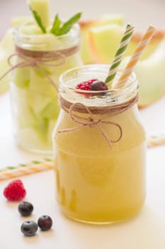 In front a jar with melon smoothie near berries, straws diagonally behind a jar with melon pieces, melon cut slices on a white background. Melon smoothie and salad in 2 jars. Vertical. Close-up.
