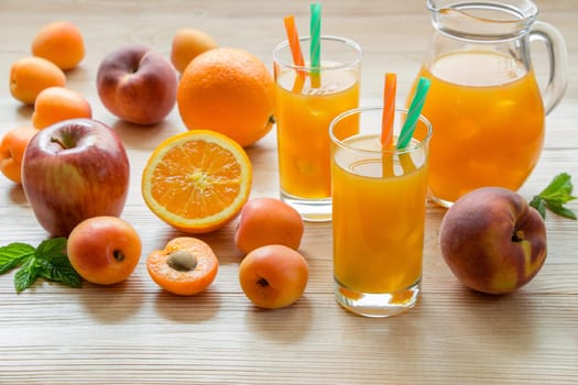 Fruit juice in glasses with ice near the jar of juice and scattered apricots, peaches, apple, orange on a light wooden background. Apricot peach apple orange juice with ice. Horizontal.