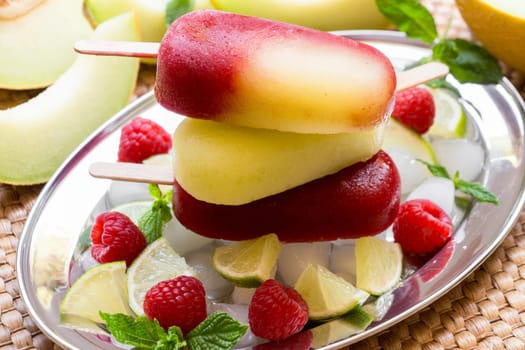 Vibrant melon raspberry ice cream sits atop a mound of ice cubes, surrounded by fresh raspberries, mint leaves, and sliced lime.