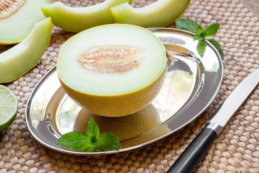 Diagonally at the center on a silver platter half of melon near melon slices, mint leaves, knife on a straw napkin background. Half of melon on silver platter. Horizontal. Close.