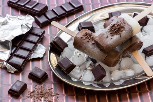 Chocolate vanilla ice cream on a stick on a silver platter with ice and pieces of chocolate near pieces of chocolate on a chocolate background. Chocolate vanilla ice cream popsicles. Horizontal.