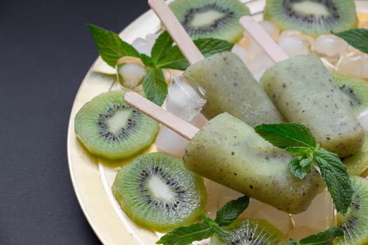 Chilled kiwi icecream popsicles are presented on a golden plate surrounded by ice cubes and fresh kiwi slices, creating a refreshing summer delight at a gathering.