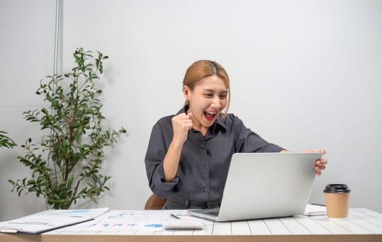 A businesswoman expresses joy and success at work, seated at a desk with a laptop, documents, and coffee in a modern office setting.