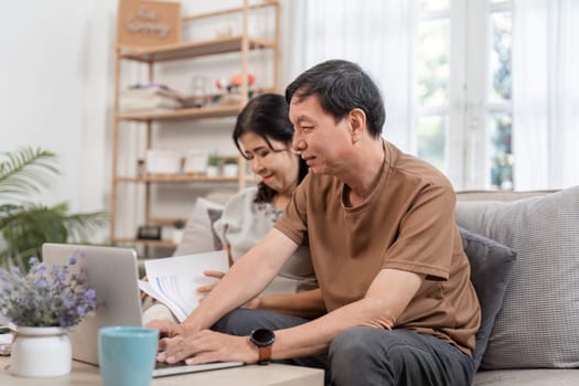 An elderly couple sits together, calculating family income and expenses using a laptop and documents in a cozy home setting.