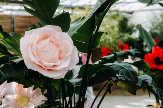 Beautiful view of giant artificial roses, gerberas and poppies made of organza in pots with real plants, which stand in a covered glass room in a public park on a sunny spring day, side view very close-up. Conceptual flower backgrounds, interior floristry, handmade hobby.