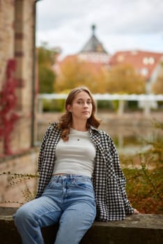 A stylish young woman with wavy brown hair and blue eyes sits on a stone ledge in a historic European city park, wearing a trendy black-and-white plaid shirt, a white crop top, and classic blue jeans. The soft natural lighting highlights the warm autumn atmosphere with golden leaves, red ivy, and medieval architecture in the background. Her relaxed pose and thoughtful expression create a cozy and nostalgic mood, perfect for lifestyle branding, travel promotions, autumn fashion editorials, and advertising seasonal collections.Young woman in casual autumn outfit relaxing in a historic European city park with medieval architecture in the background.