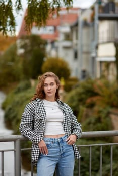 Young woman in trendy autumn outfit posing on a European bridge with golden foliage and a historic cityscape in the background. A stylish young woman with wavy brown hair and blue eyes poses confidently on a bridge in a European city, surrounded by vibrant autumn foliage. She wears a black-and-white plaid shirt over a white crop top, paired with blue jeans and white sneakers, creating a fashionable yet casual look. The river flowing beneath the bridge, the historic architecture in the background, and the golden tones of the fall season add depth and warmth to the scene. This high-quality image is ideal for lifestyle branding, autumn fashion campaigns, travel blogs, social media content, and seasonal advertising.