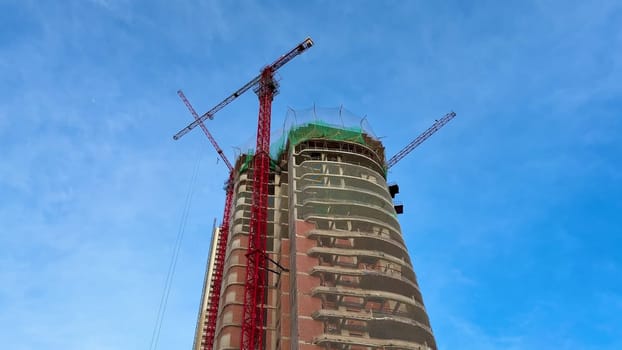 The construction process of a tall, modern skyscraper is underway, featuring cranes against a clear blue sky