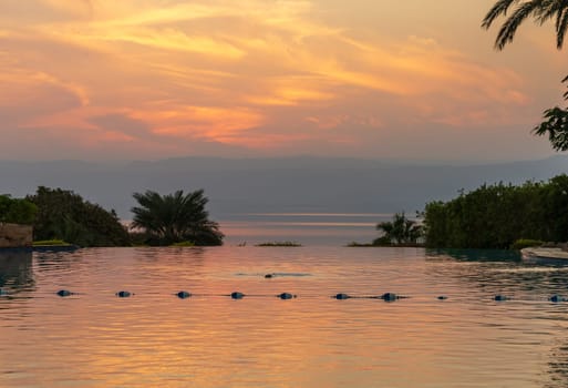 Flowers and palm tree frame the sunset over wimming pool and the Dead Sea in Jordan with Israel in the distance