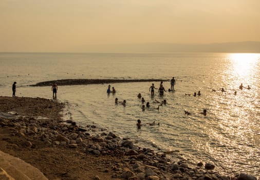 Tourists in the waters of Dead Sea by a beach in modern hotel at sunset