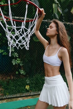 Athletic young model showcasing confidence and style next to a basketball hoop, wearing a white crop top and skirt, depicting themes of fitness and sports