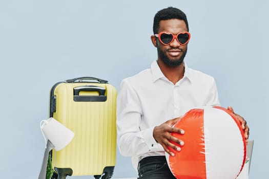 Smiling man with dark skin and glasses, dressed in white shirt, holding inflatable beach ball, standing next to yellow suitcase, studio shot against plain light blue background, summery travel, holiday mood, carefree lifestyle, vacation concept, leisure.