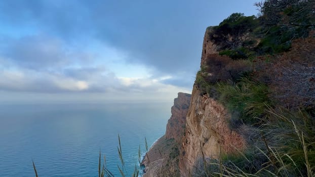 A breathtaking view of a coastal cliff with lush greenery and tranquil ocean waters under a cloudy sky.