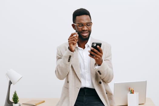 Cheerful man with dark skin and glasses interacting with smartphone in clean, minimalist office setting, dressed in casual business attire, with laptop and plant on desk, conveying modern work communication, positive mood. Concept: digital communication, modern workspace