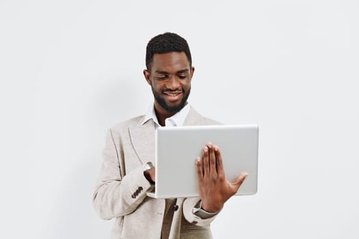 Smiling young man with dark skin, dressed in a light-colored suit, holding a laptop and engaging with the device in front of a plain white background. Concept: professional work, technology, confidence, success, corporate lifestyle.