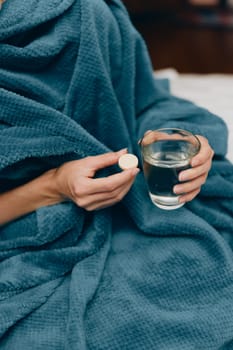 Woman in blue blanket sitting on bed, holding glass of water and pill in hand.