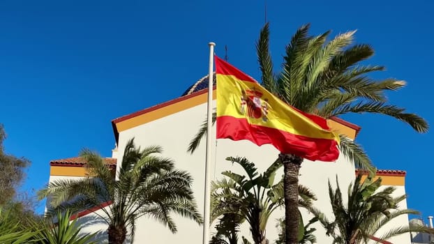 A vibrant Spanish flag waves in the breeze, set against a bright blue sky and lush green palm trees swaying