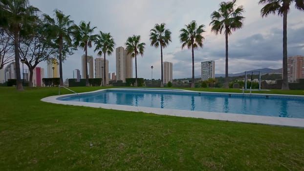A scenic poolside scene surrounded by palm trees and contemporary city buildings under a cloudy sky.