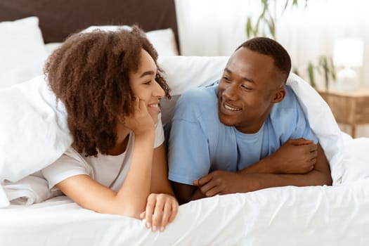 Enjoy communication with each other on weekends and relax at home during covid-19 quarantine. Cheerful millennial african american couple under covers, woman listen male in bedroom, copy space