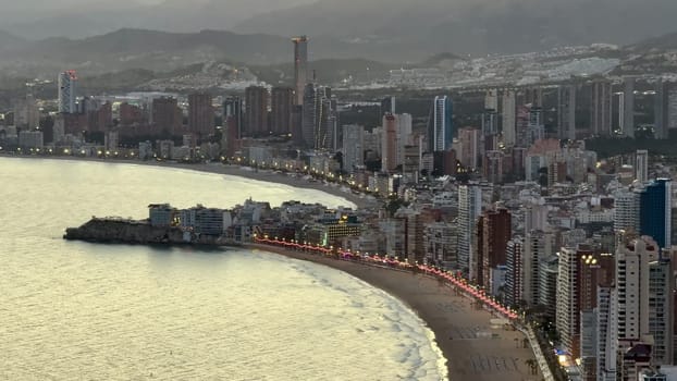 A picturesque view of a modern coastal city with illuminated skyline and tranquil waters during sunset.
