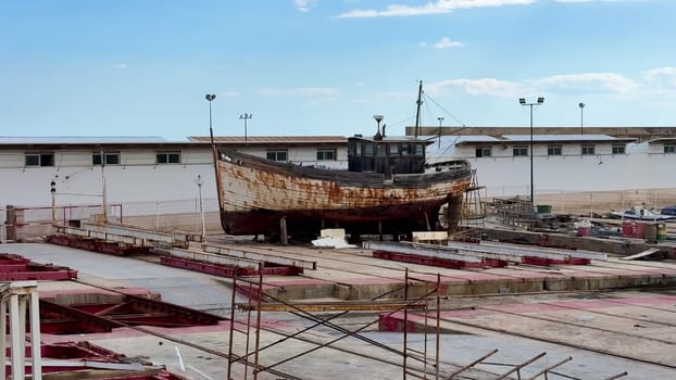 An old, rusted fishing boat is docked in a desolate shipyard, all under a bright blue sky that adds to the scene
