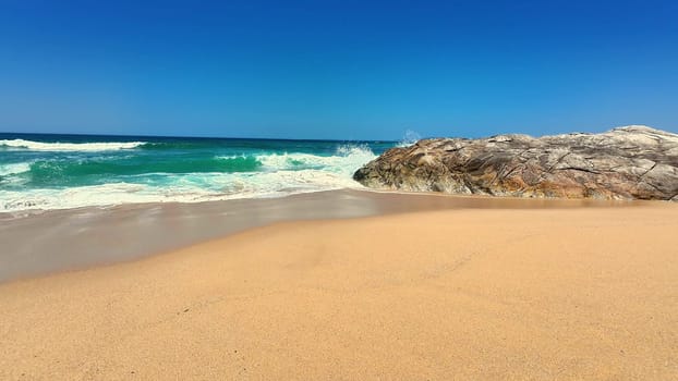A picturesque beach scene featuring soft sand, gentle waves, and a large rock formation under a clear sky.