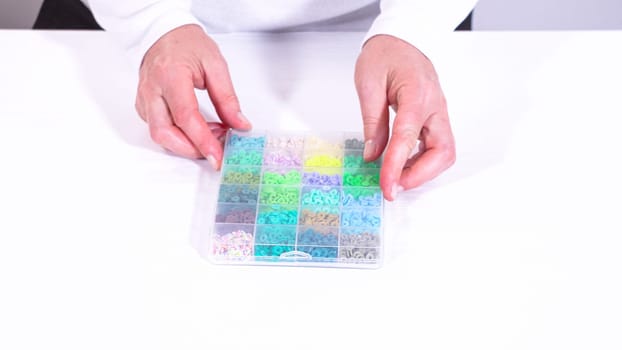 Womans hands gracefully poised over a collection of beads, sorted by color in transparent organizers. The array of beads spans a vibrant spectrum, from deep purples to bright oranges, meticulously arranged for easy selection as she embarks on creating a custom piece of jewelry.