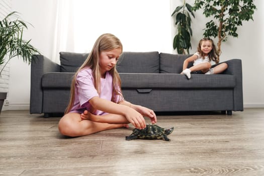 Two children interact in a bright living room. One girl sits on the floor, gently petting a turtle, while the other watches from the couch, adding a lively atmosphere.