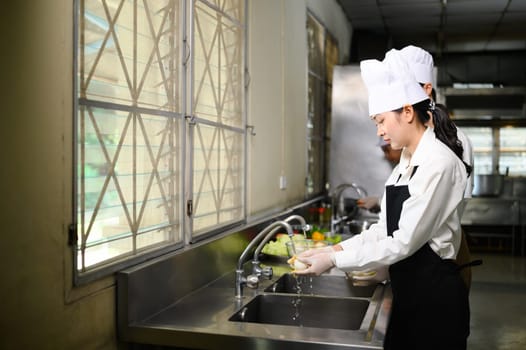 Young chef in uniform washing vegetables at the sink during culinary class.