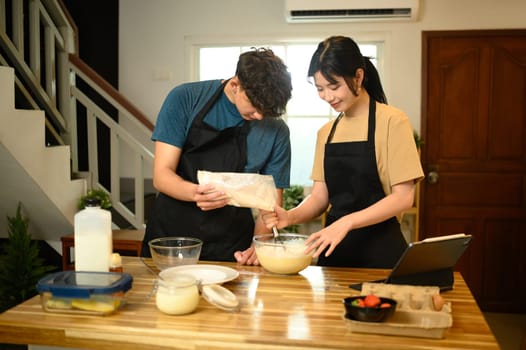 Man and woman cooking breakfast at home following digital recipe to make pancakes in a kitchen.