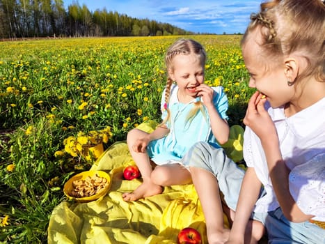 Two young girls share laughter and snacks on a blanket in a colorful dandelion field under a clear blue sky.