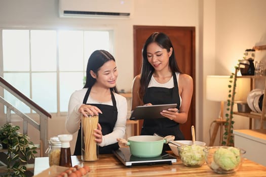 Asian mother and daughter cooking together in kitchen and looking recipe on digital tablet.