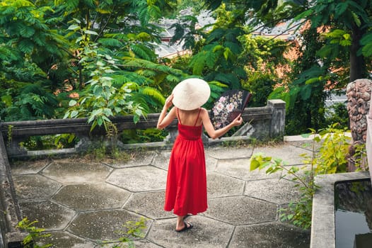 back of beautiful woman tourist traveler in a Vietnamese non la hat with a fan in a garden in Vietnam. Tourism and travel in Asia