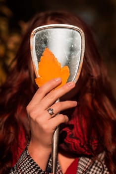 young girl holding yellow autumn leaves in her hands against the background of a mirror. autumn concept. she is wearing a coat. her face is not visible.