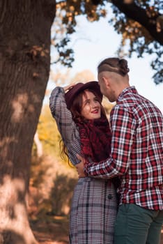young couple in love walking and having fun in autumn park. they look at each other and hug, against the background of autumn trees. the girl is wearing a dark red hat. concept of romance