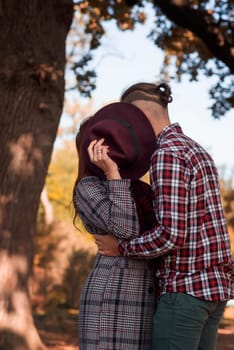 young couple in love hugging and kissing in autumn park, covering themselves with hat. girl in coat, against autumn trees, golden autumn around. concept of falling in love, relaxation