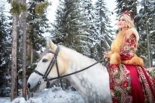 A stunning girl dressed in a vibrant national outfit rides a horse, celebrating Maslenitsa in a snowy forest setting.