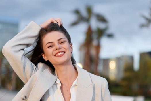Confident young woman in beige business jacket smiling outdoors with urban cityscape and palm trees blurred in background, natural light enhancing fresh and professional business vibe