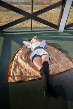 A woman lies comfortably on a blanket in a park enjoying sunny weather while wearing a stylish outfit, relaxing under a clear sky.