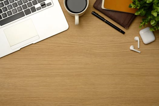 Top view of a wooden desk with laptop, coffee, notebook and wireless earbuds..