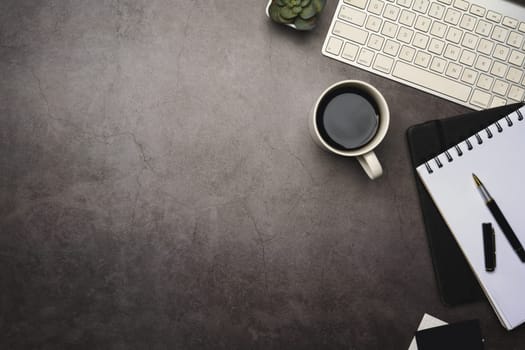 Flat lay of office desk with keyboard, coffee cup and notebook on dark stone surface.